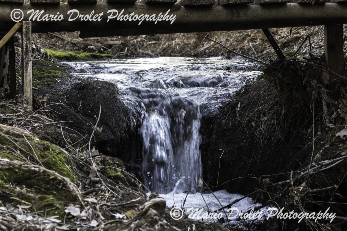 Colour photo of a small creek waterfall under a wooden bridge